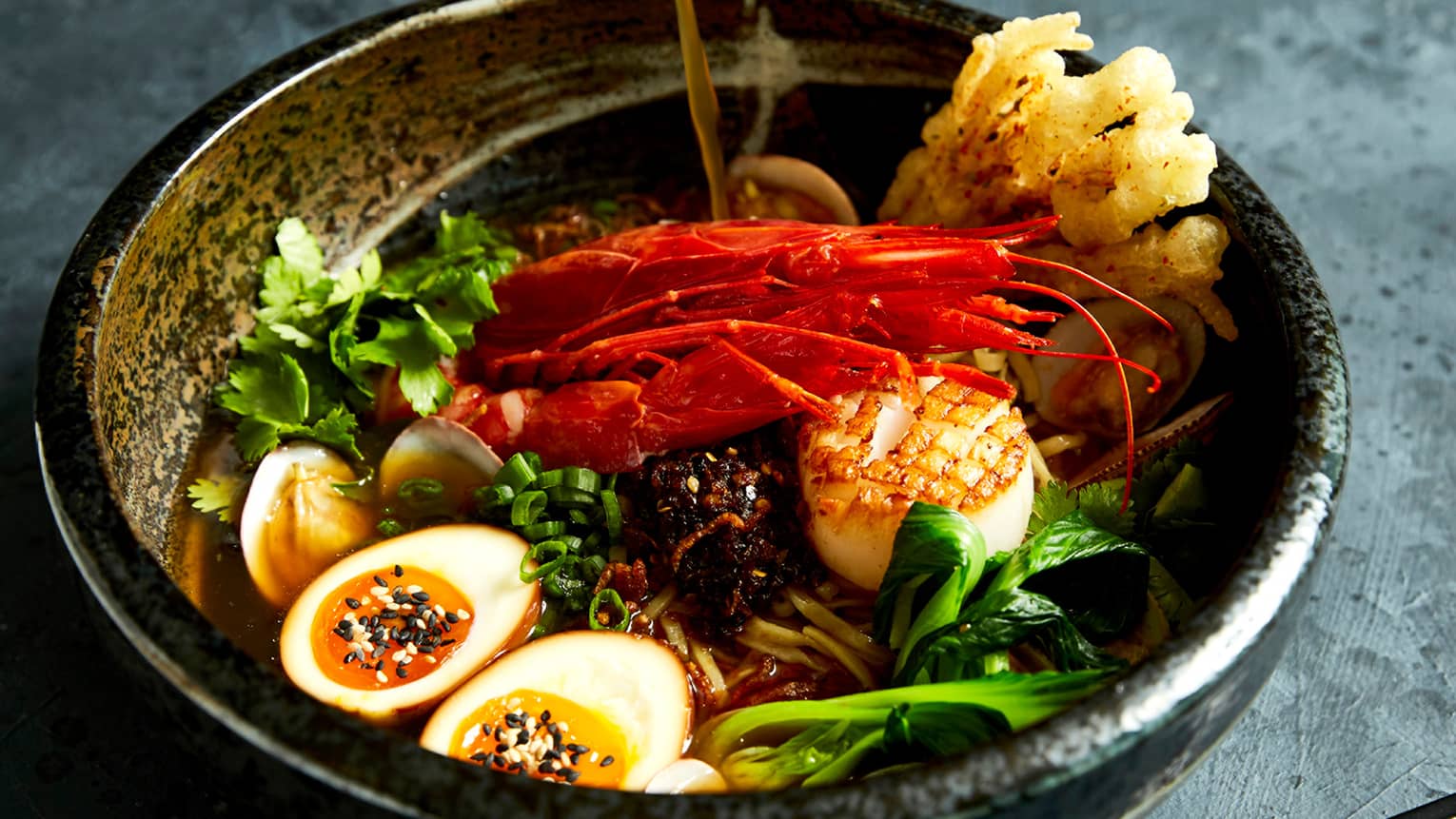 A brass bowl holding poached eggs, lobster claws, scallions and greens