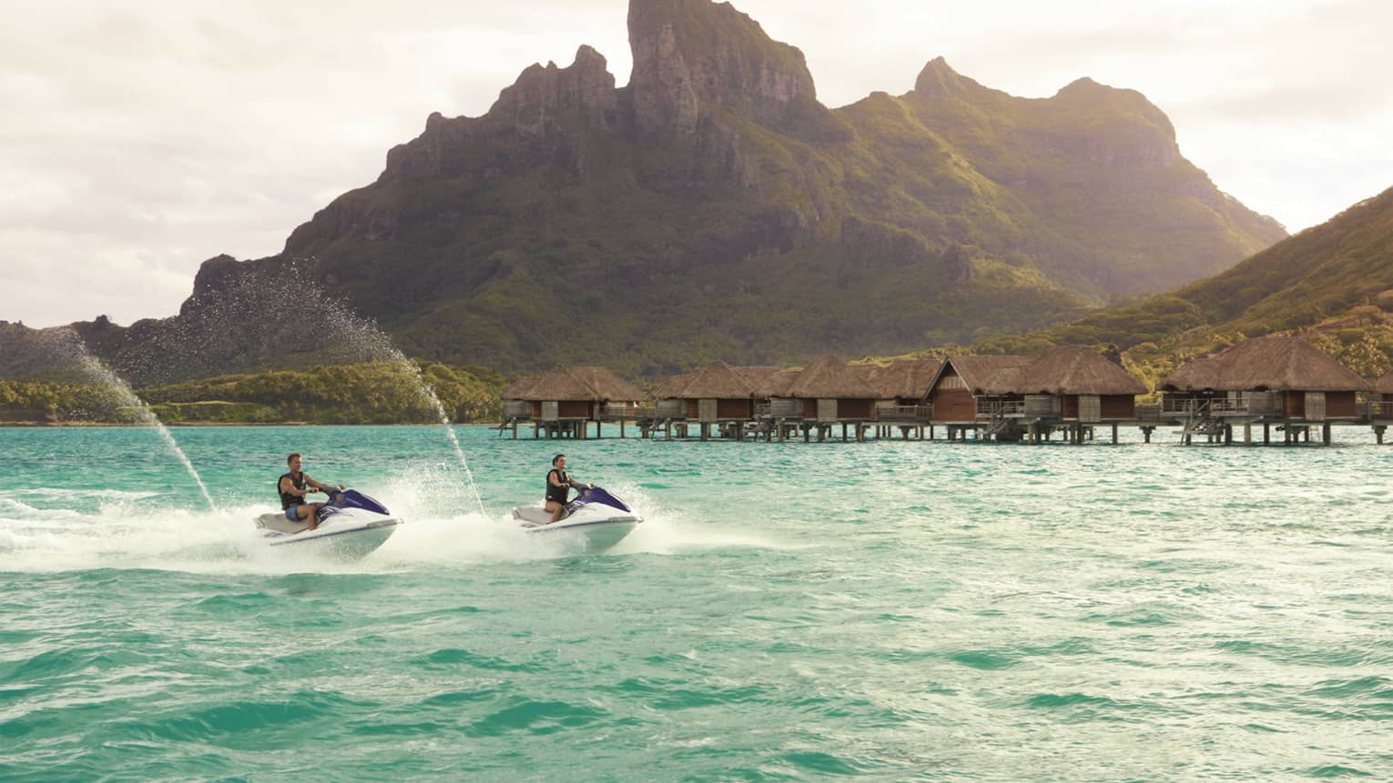 Two jet skis ride along lagoon past row of overwater bungalows, Bora Bora mountain