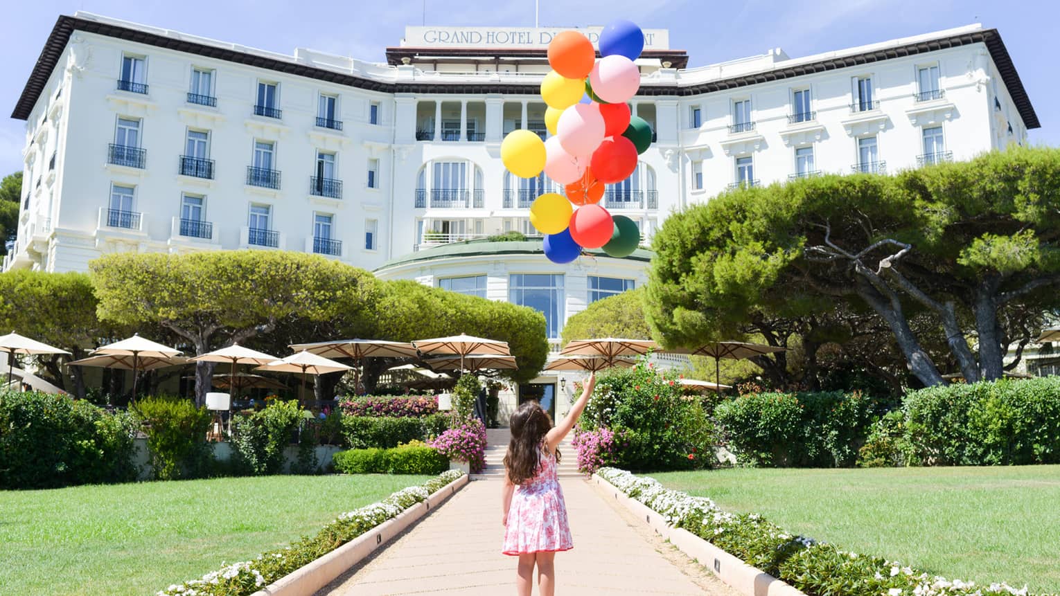 Young girl holds bunch of colourful balloons on brick path leading to hotel