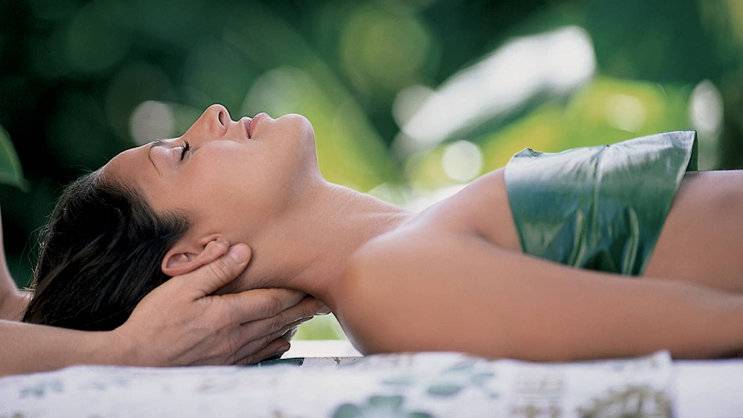 Woman with eyes closed lays on back on table with leaf over chest, hands massage her neck