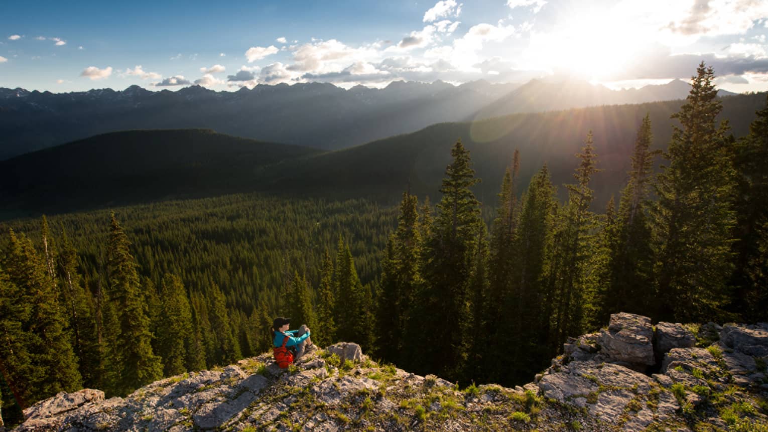 Person sits at edge of cliff overlooking trees, mountains as sun starts to set