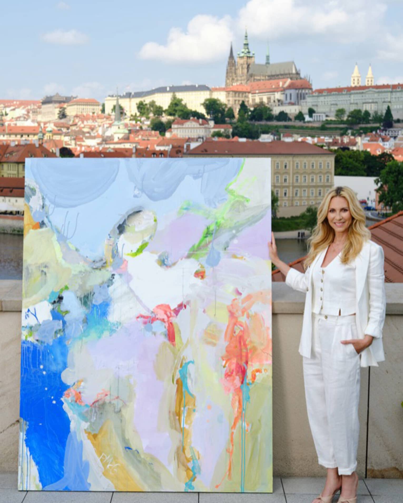 Artist Petra McGrath stands next to a colorful painting on a rooftop overlooking the city of Prague