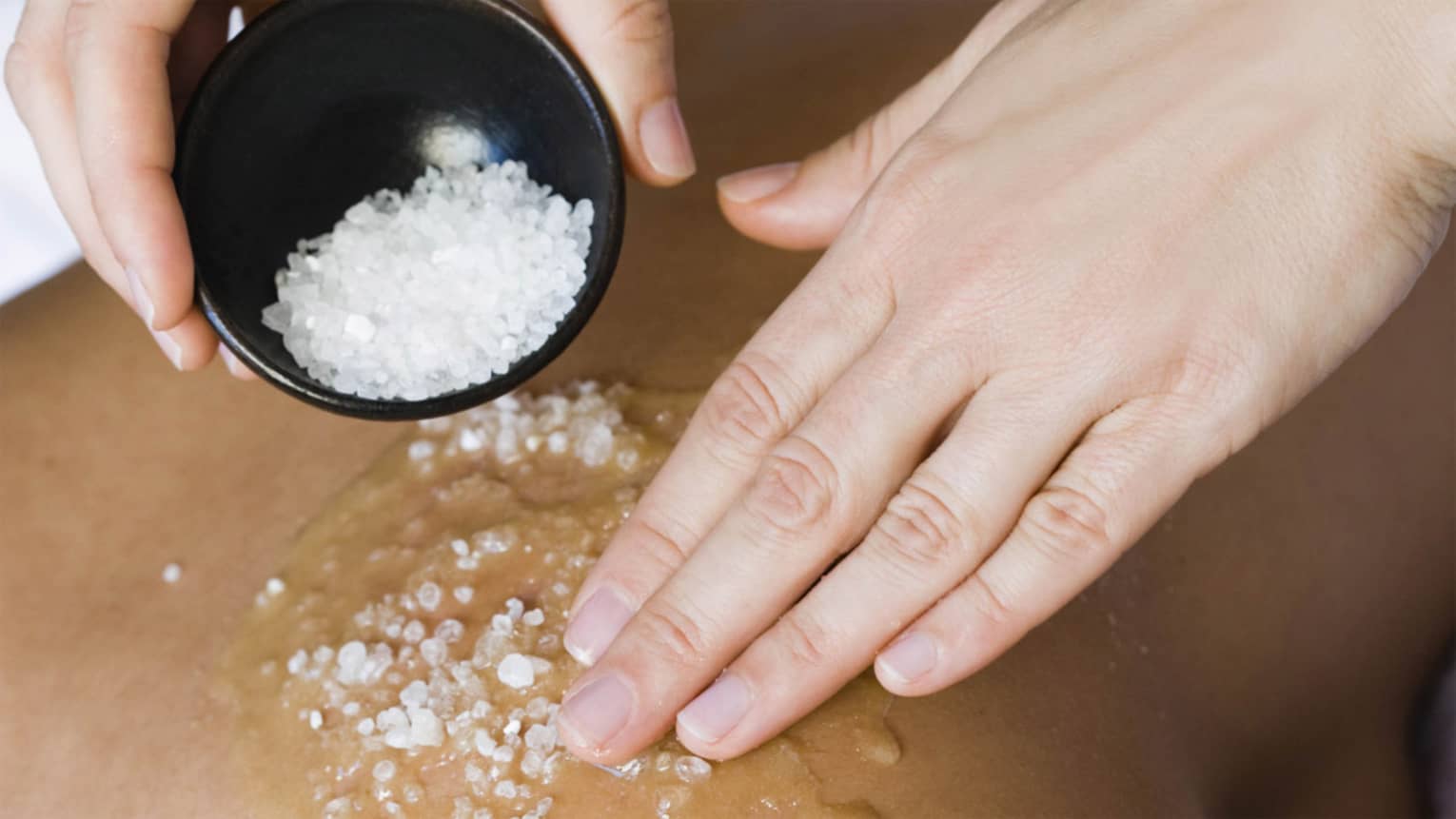 Close-up of large spa salts in bowl, hand rubbing crystals on bare back