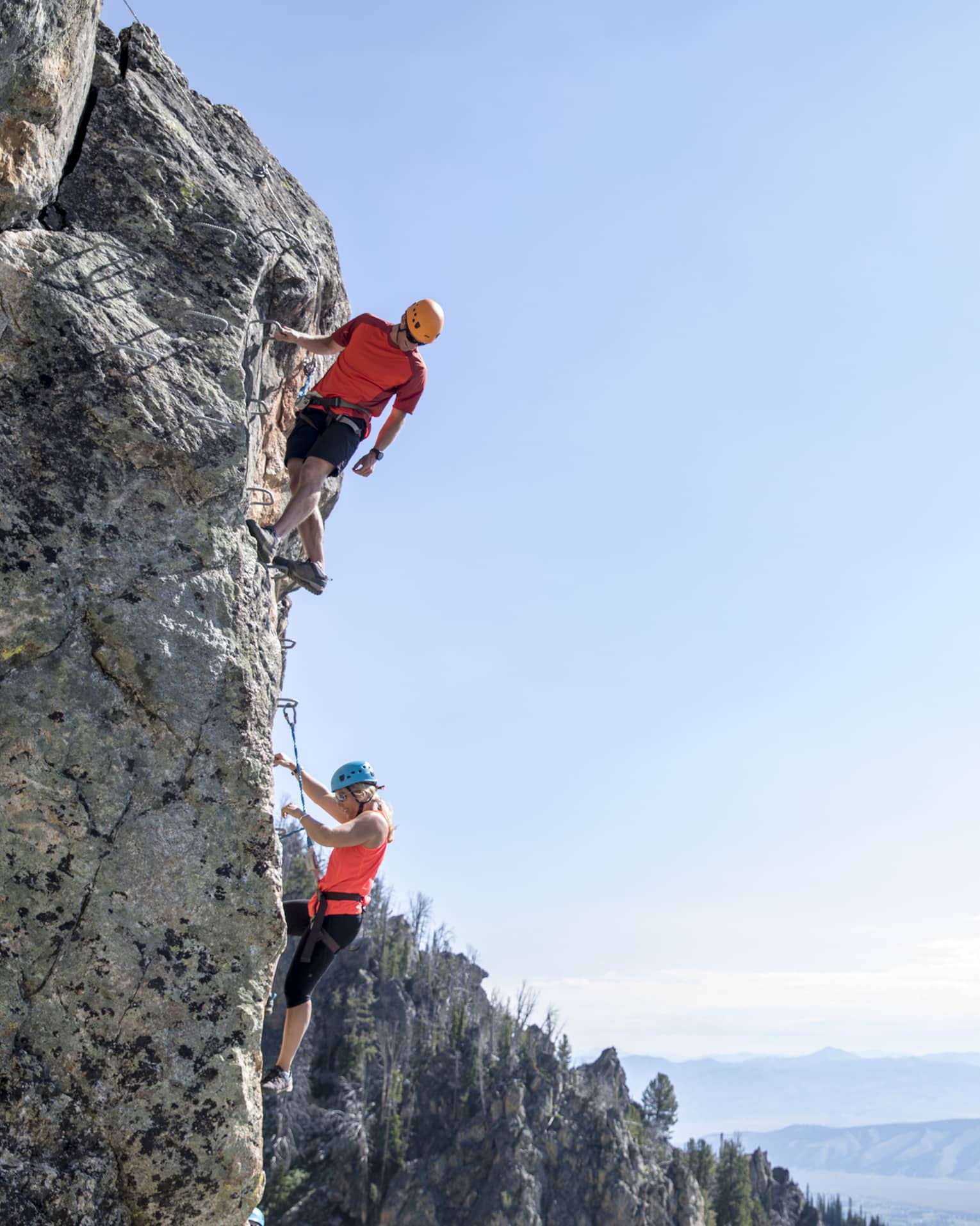 Man and woman climb steep cliff face with view of mountains in far distance 