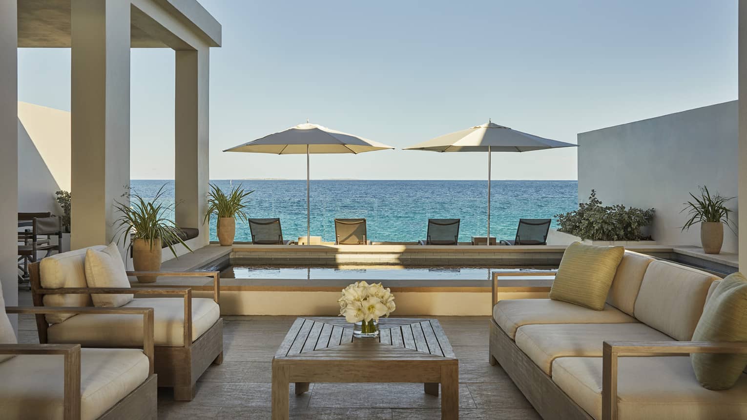 Outdoor patio living room with sofas, armchairs with white cushions, plunge pool, patio chairs and umbrellas, ocean