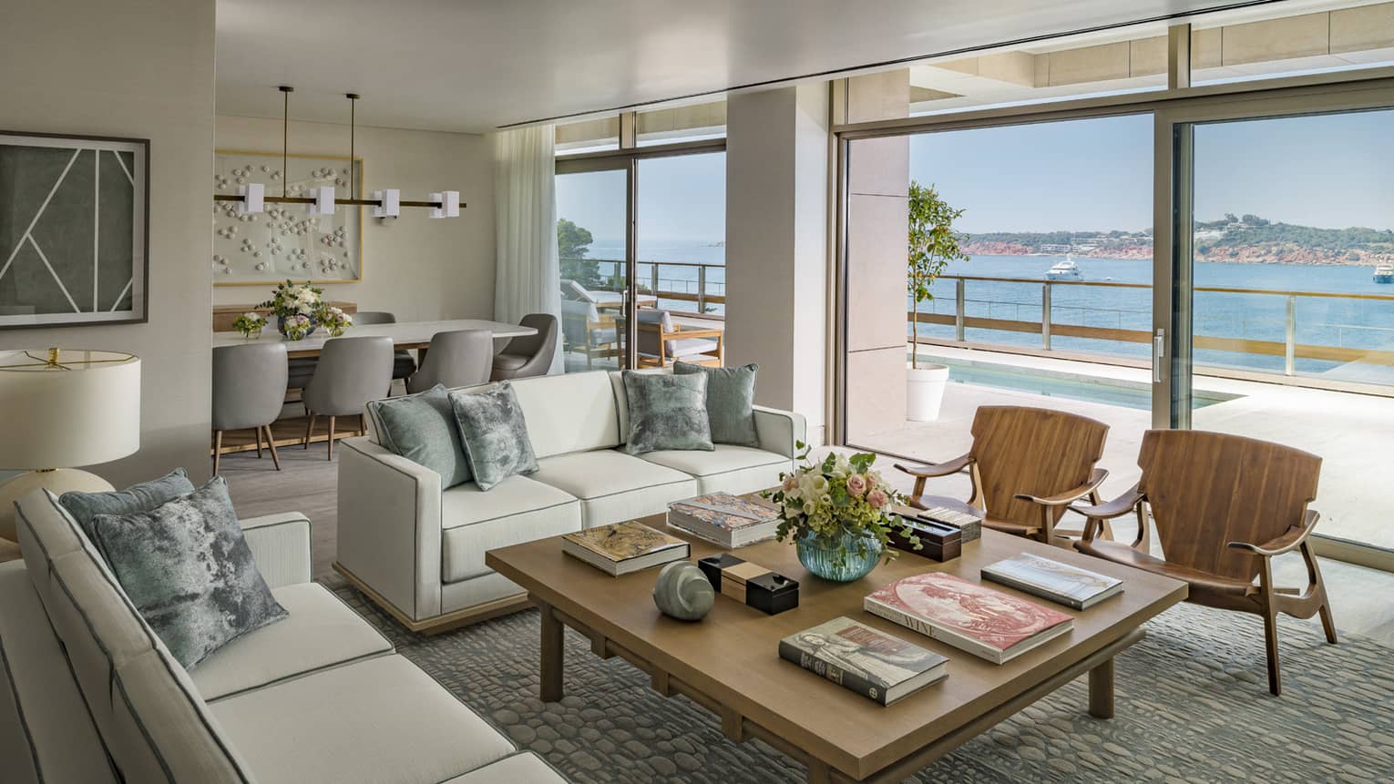 Arion Royal Suite living area with white sofas, square coffee table, arm chairs, dining table in background, floor-to-ceiling window with view of plunge pool and Mediterranean Sea