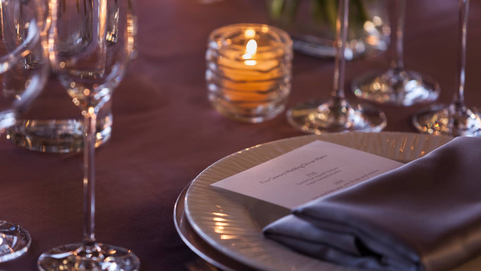 Close up of table with candle, grey napkin and dinner menu on white plate, glasses 