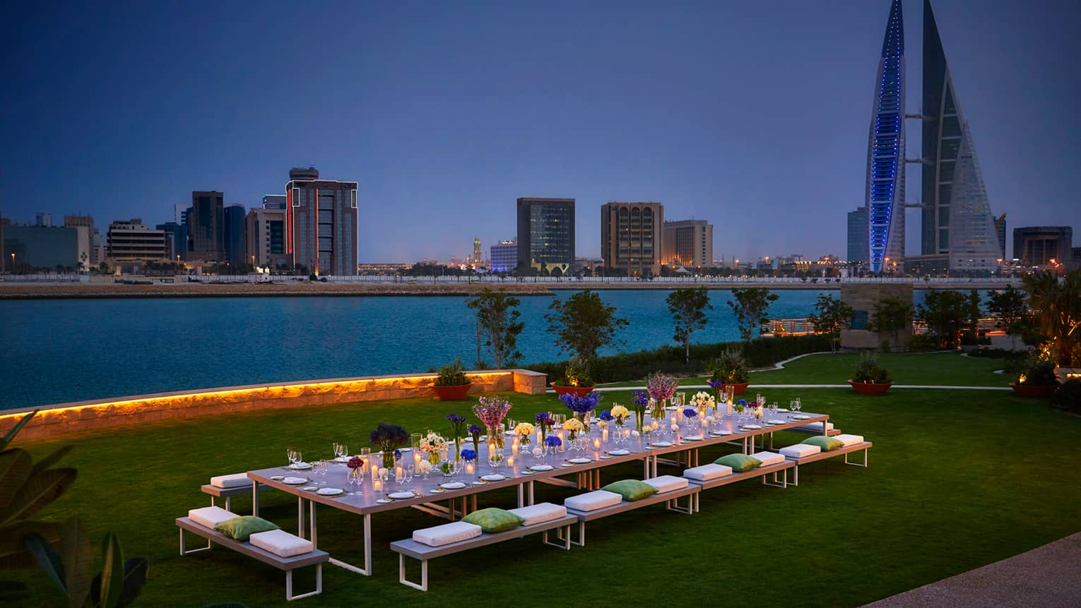 Long, candle-lit dining table on event lawn by water at night