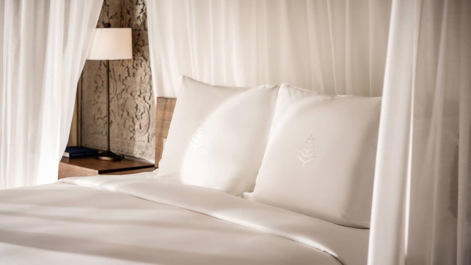 Close-up of bed headboard, white pillows with Four Seasons logo embroidered, crisp white linens, white curtains