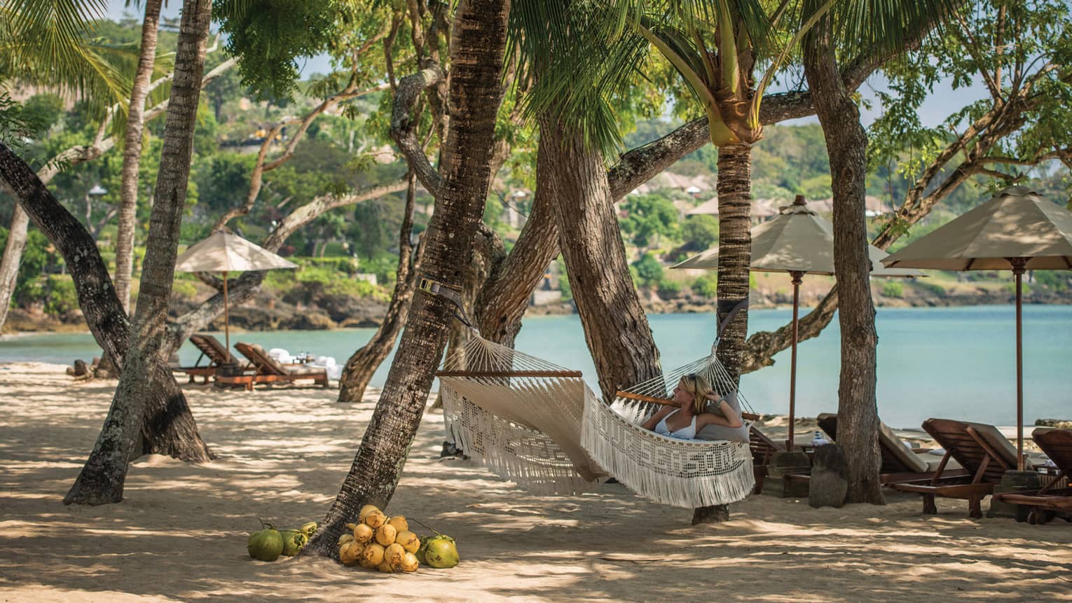 Woman in white bikini lies on white hammock in shade of large trees on Coconut Grove Beach