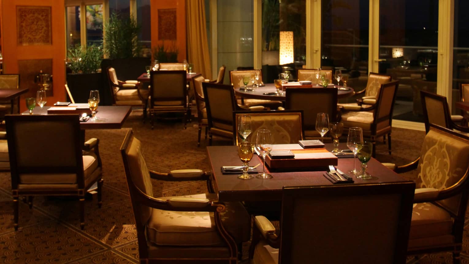 Dimly lit dining room of restaurant, brown square tables, each with four arm chairs, windows