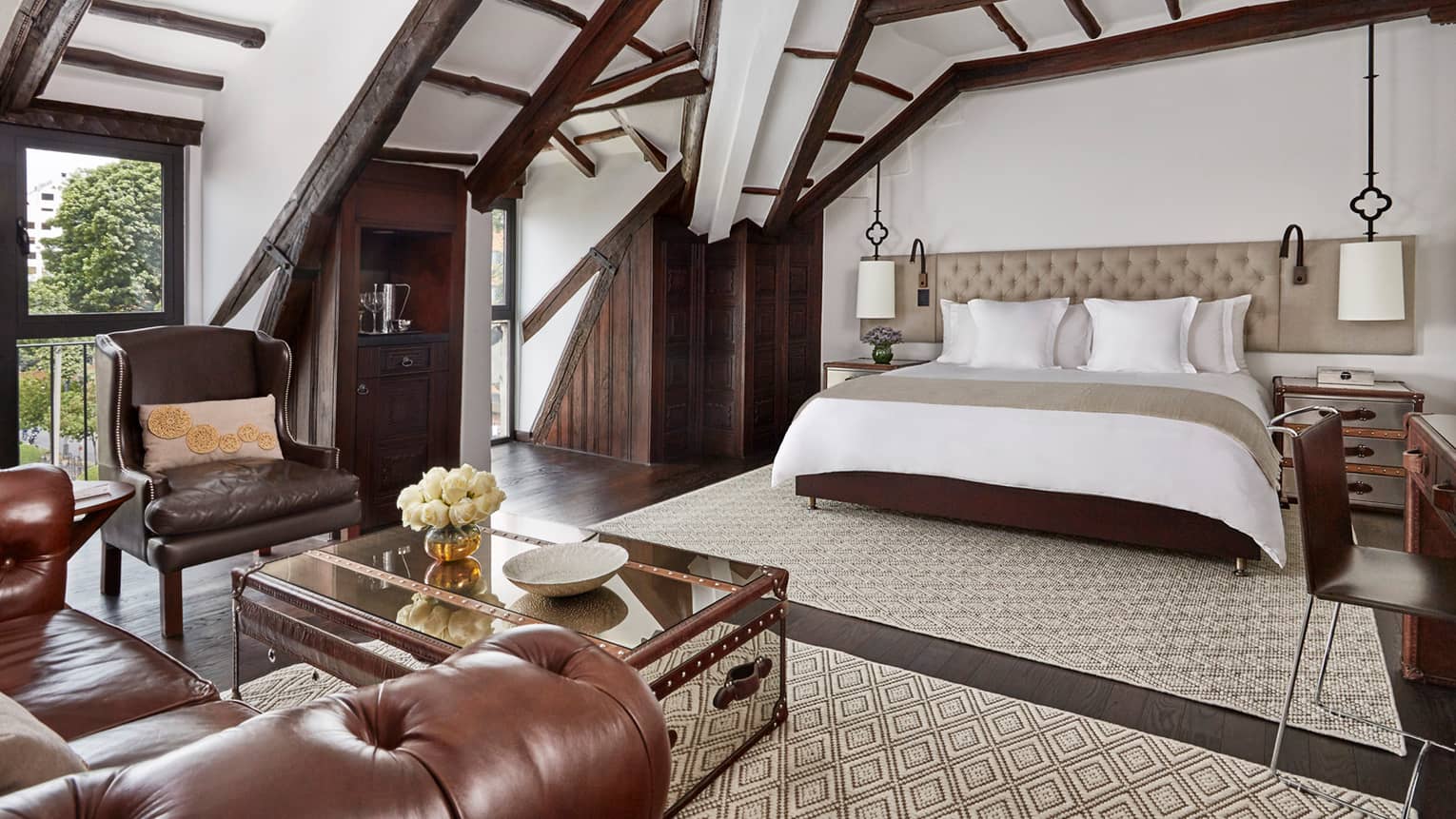 Sloped beam loft ceiling over hotel bed with padded headboard, brown leather armchair, table with flowers