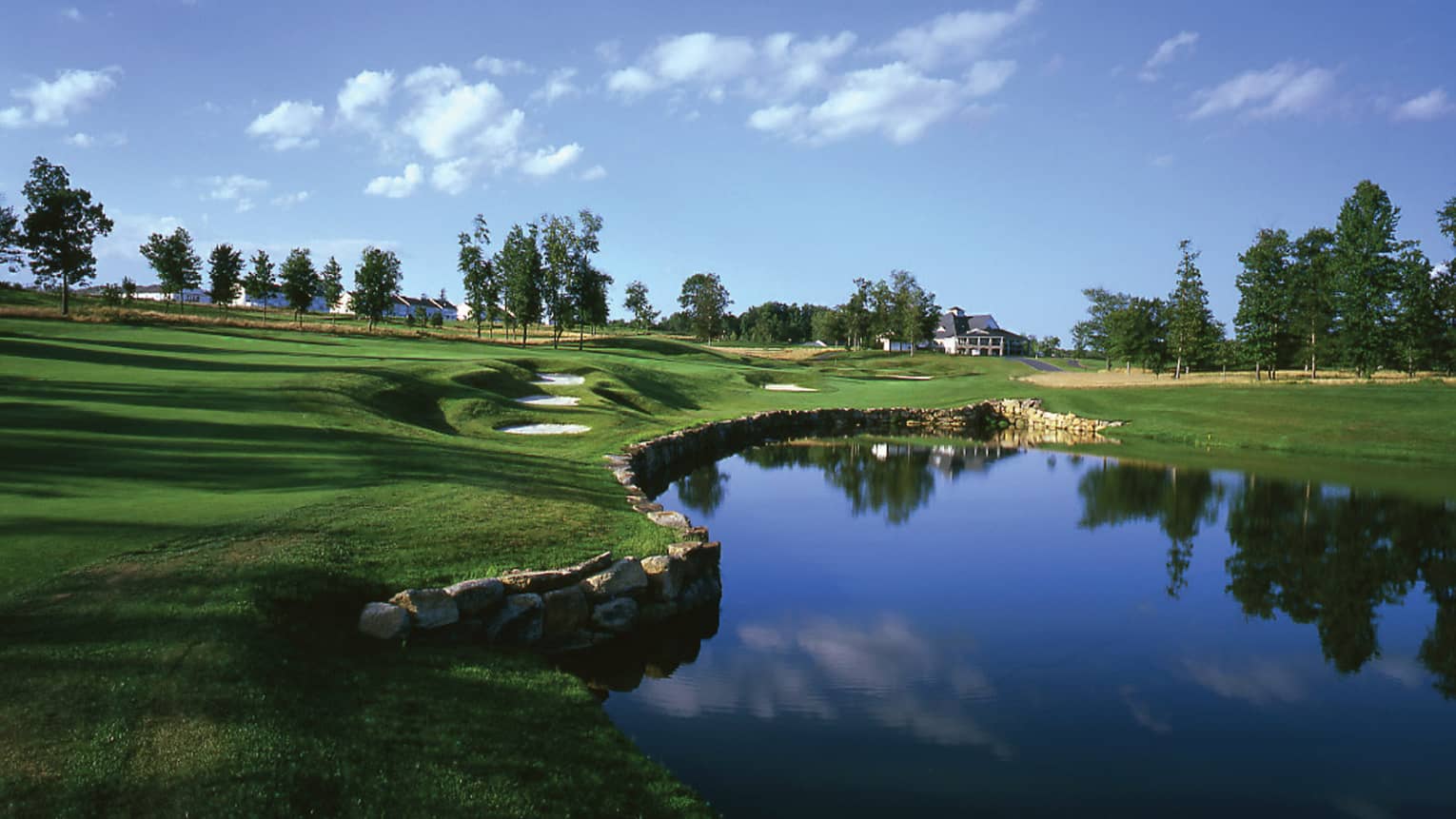 Pond in front of rolling golf course greens under blue sky