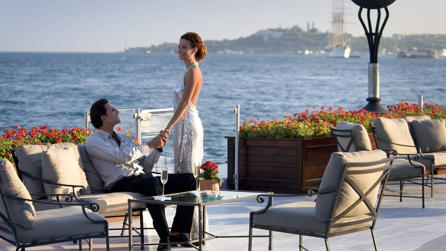 Woman stands over man on patio lounge chair by table with Champagne, water 