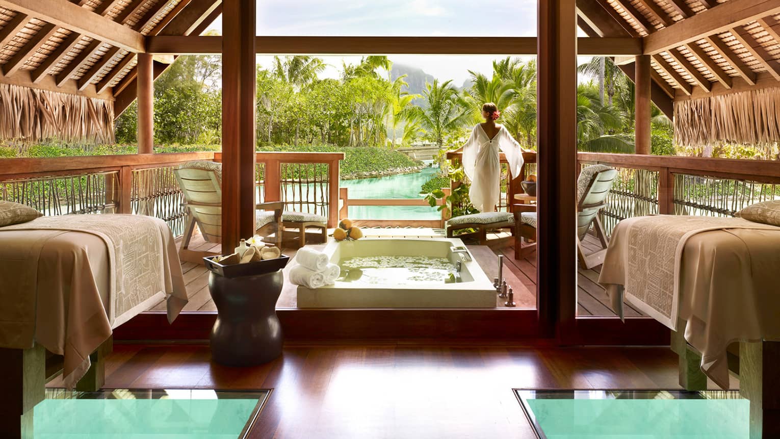 Woman wearing flowing white robe stands at edge of spa suite near plunge bath, couples massage beds