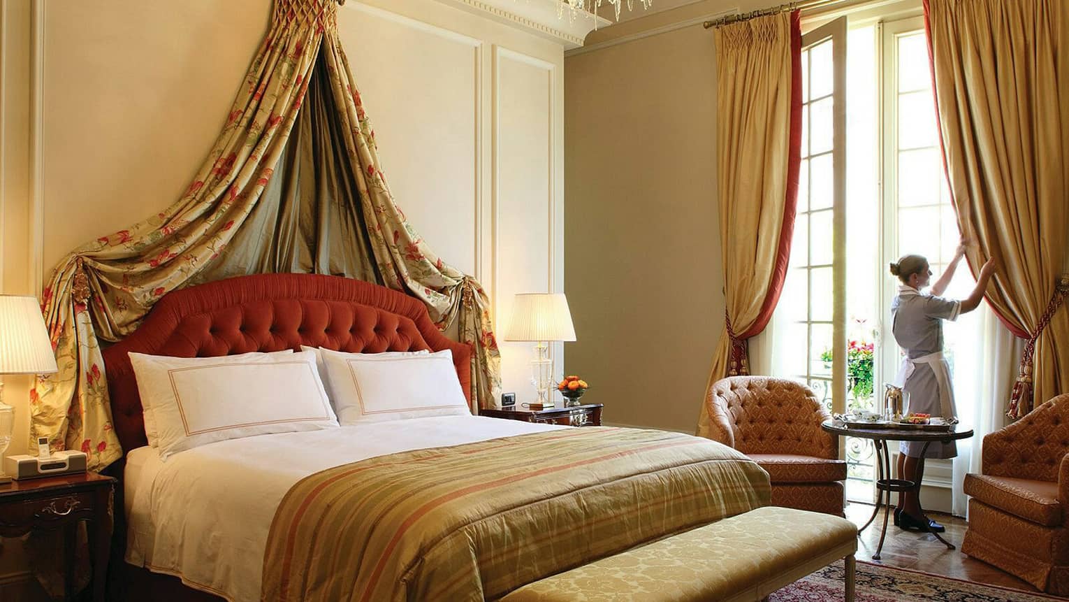 Hotel room bed with red velvet headboard with gold-and-red patterned drapes and bedspread, hotel staff opening curtains