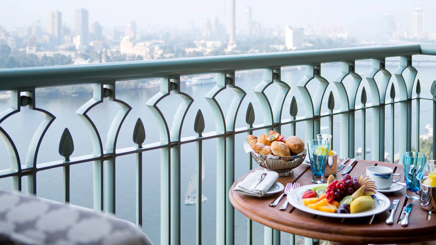 Close-up of balcony table with plate of fresh fruit, bread and orange juice, river and city view below