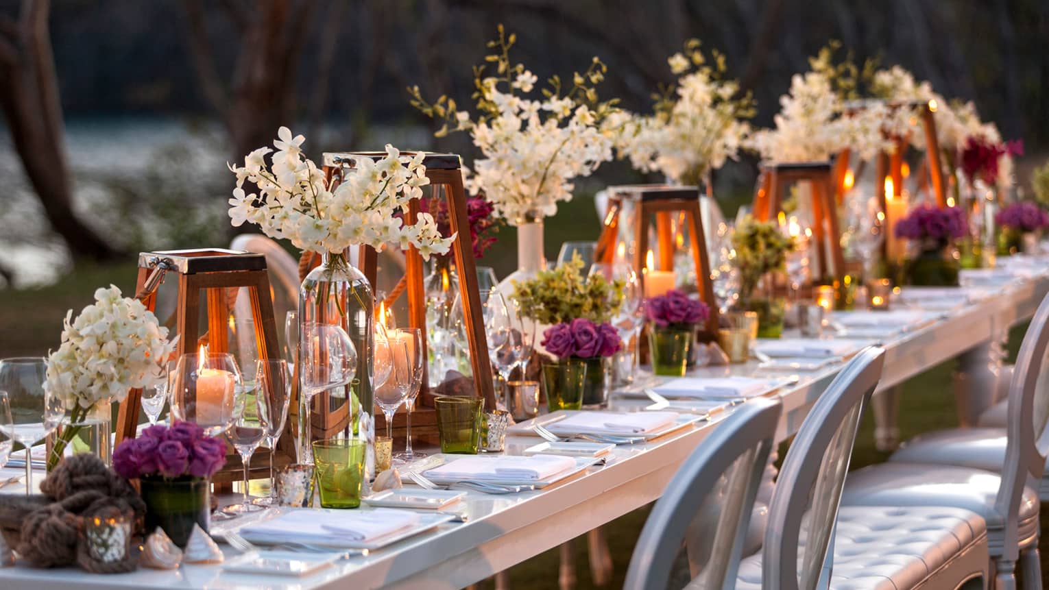 Long outdoor dining table with glowing candles in glass lanterns, white and purple flowers 