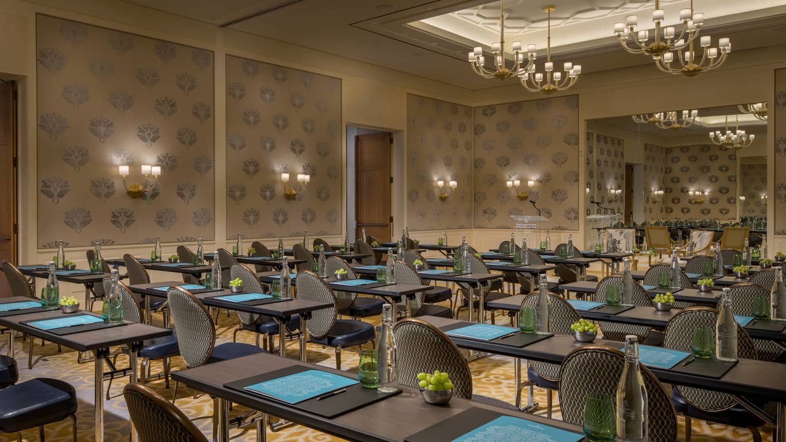 Rows of tables, chairs under high ceilings with small chandeliers in large meeting room