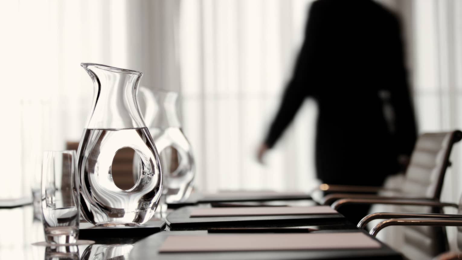 Person wearing business suit walks behind meeting table with agendas, glass water jugs