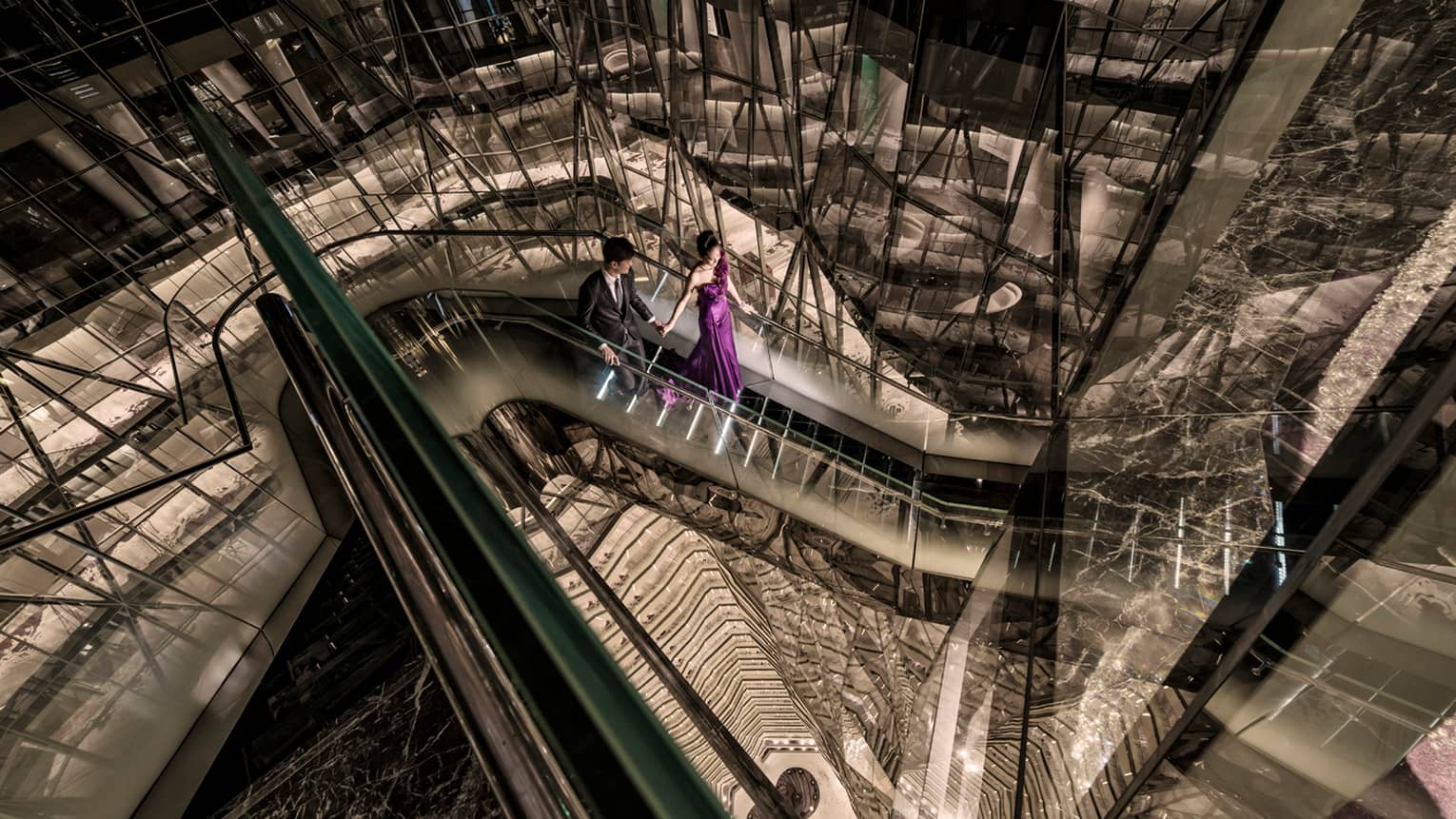 Woman in long purple evening gown and man in suit descend down glass escalator high in tower