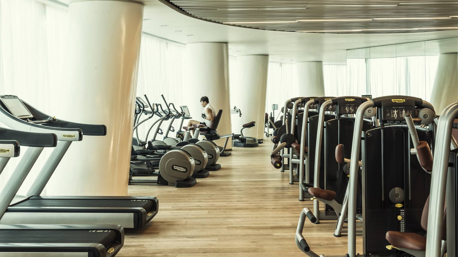 Cardio machines in bright fitness room with modern white pillars, man on bicycle machine