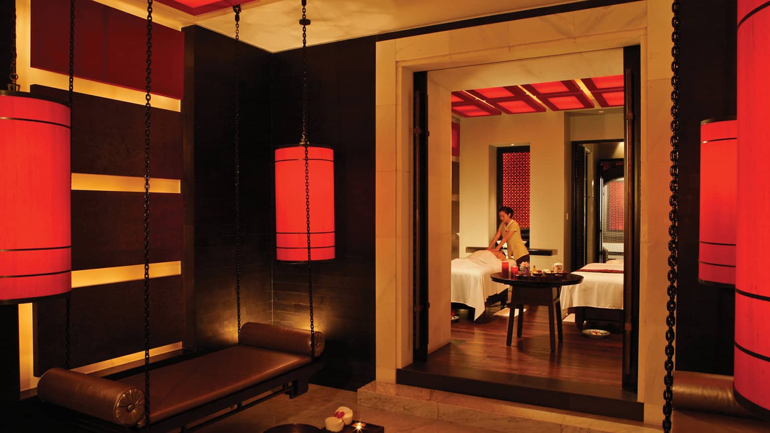 Spa staff gives massage on table behind doorway in dark spa with hanging red lanterns
