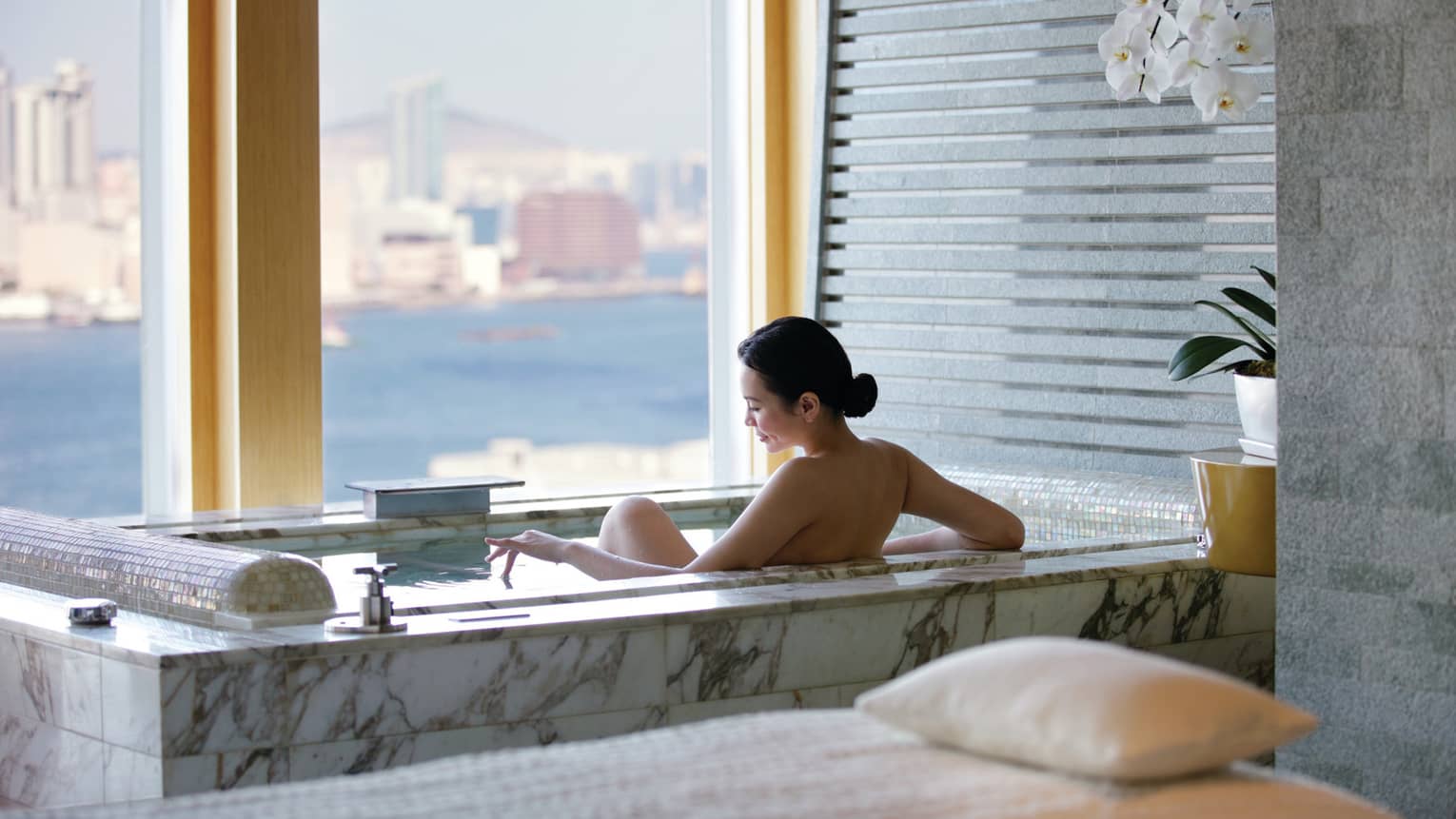 Woman's bare back above marble spa tub in tile bathroom by window with water, city views