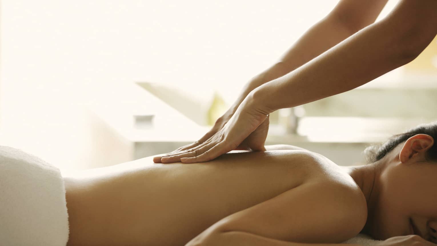 Masseuse reaches over, massages woman's bare back as she lies on spa table