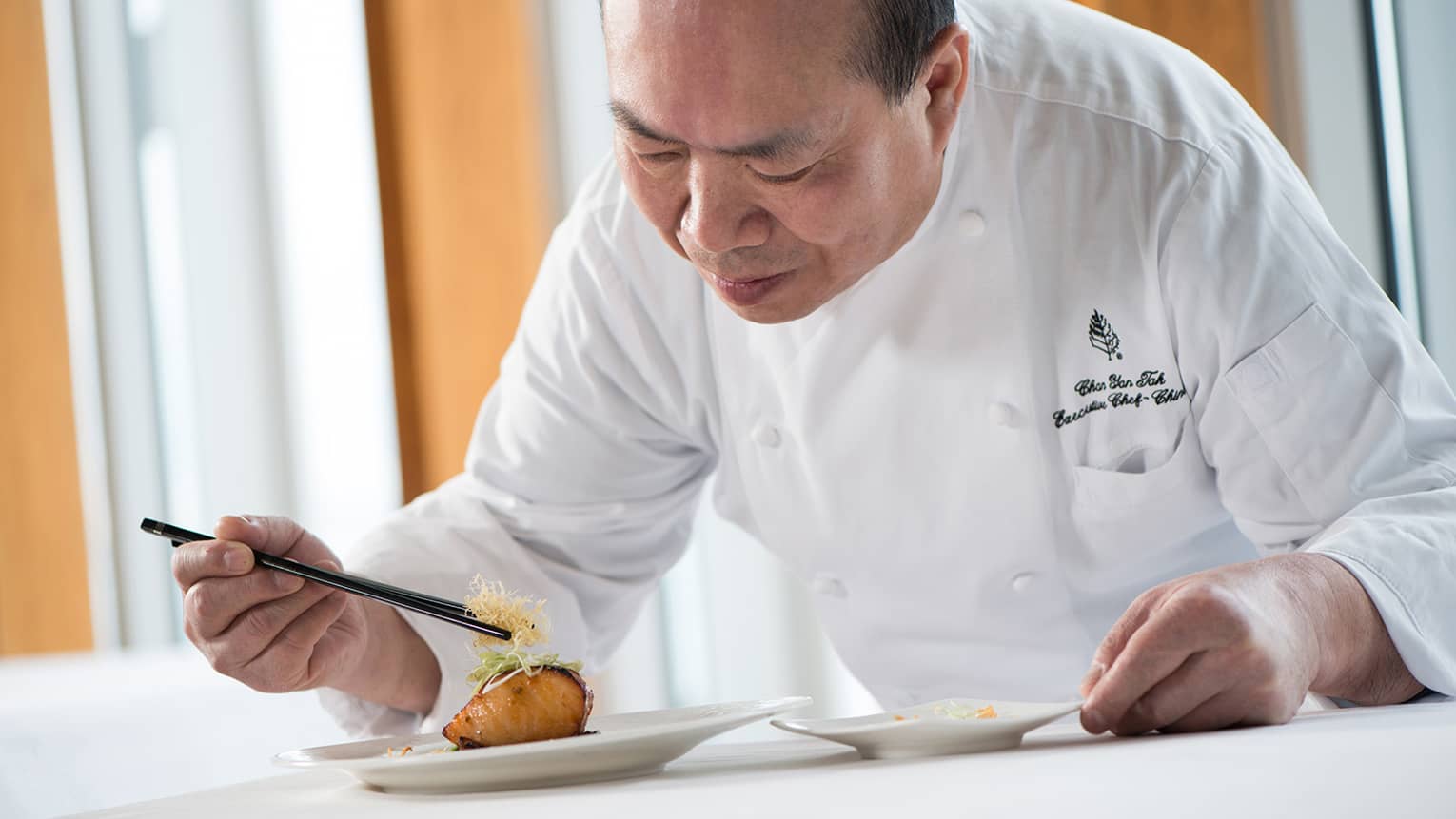 Chef in white uniform leans over grilled salmon dish on plate, adds garnish with chopsticks
