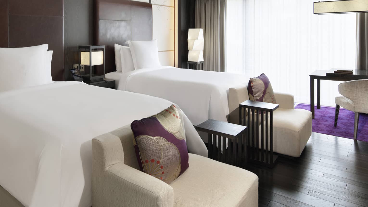 Hotel room with two twin beds, wooden floors, beige arm chairs, purple rug