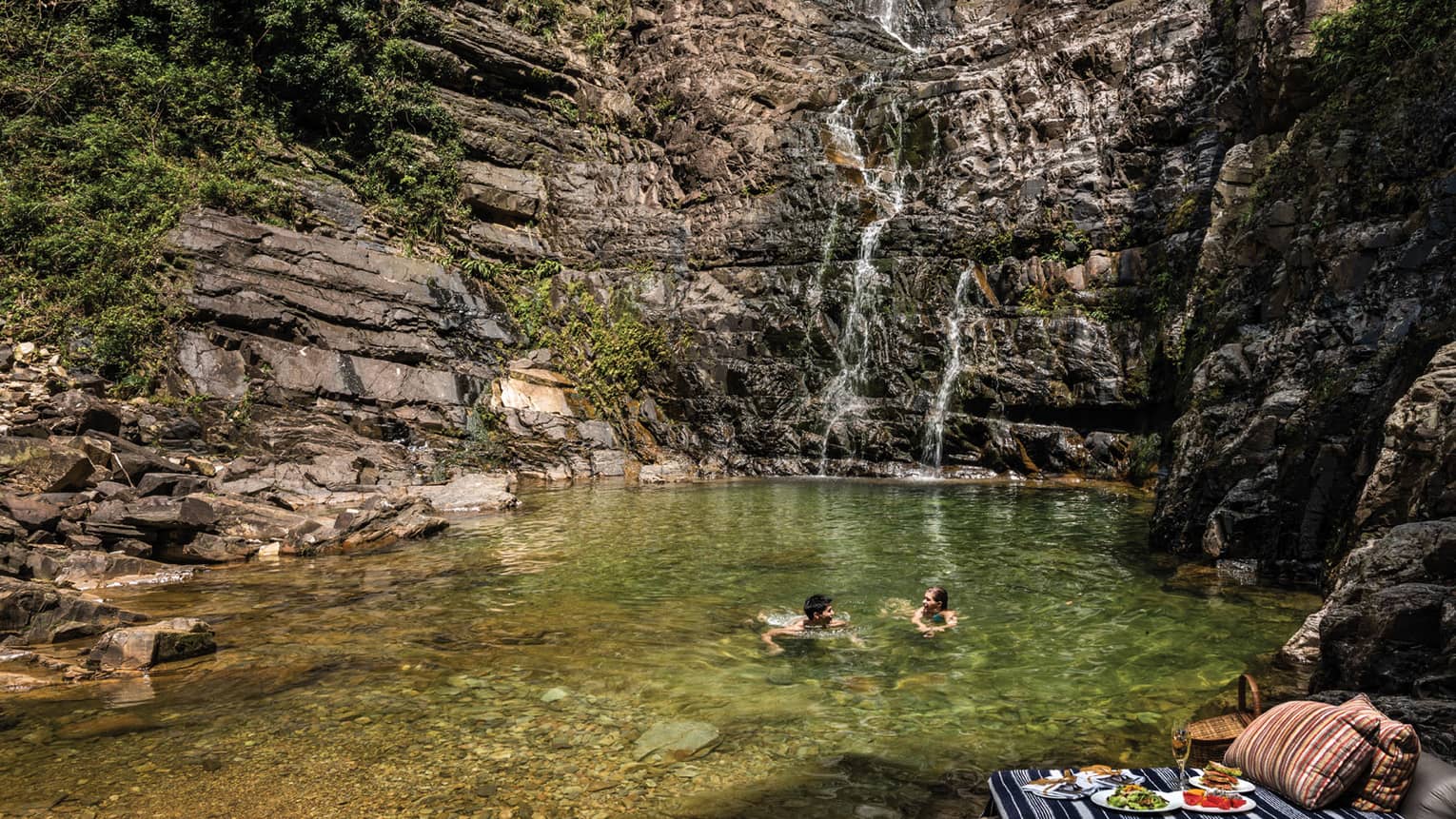 Couple swims in pool at base of waterfall, picnic lunch on rocks