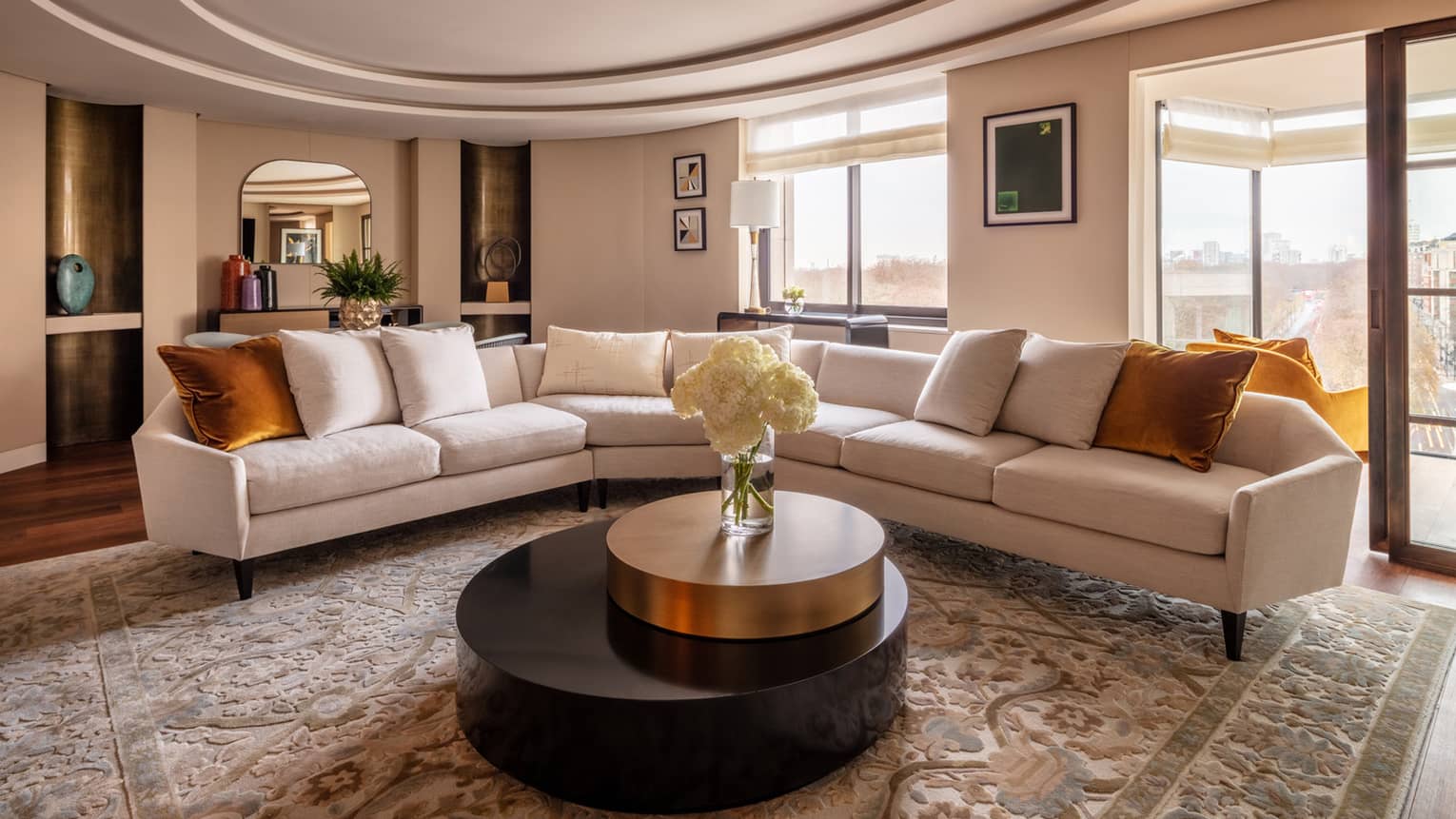 The Hyde Park Suite at Four Seasons London Park Lane in Mayfair showcases a sophisticated living room with a panoramic view of the city. The room features a long, sleek white sofas with orange throw pillows, arranged around a large circular black coffee table with a gold rim. The area is adorned with a luxurious beige patterned rug, adding texture and warmth. Artworks hang on the walls, and a large window frames the stunning view outside, providing ample natural light. The decor is elegantly completed with a neutral colour palette and modern furnishings, creating an inviting and stylish space.