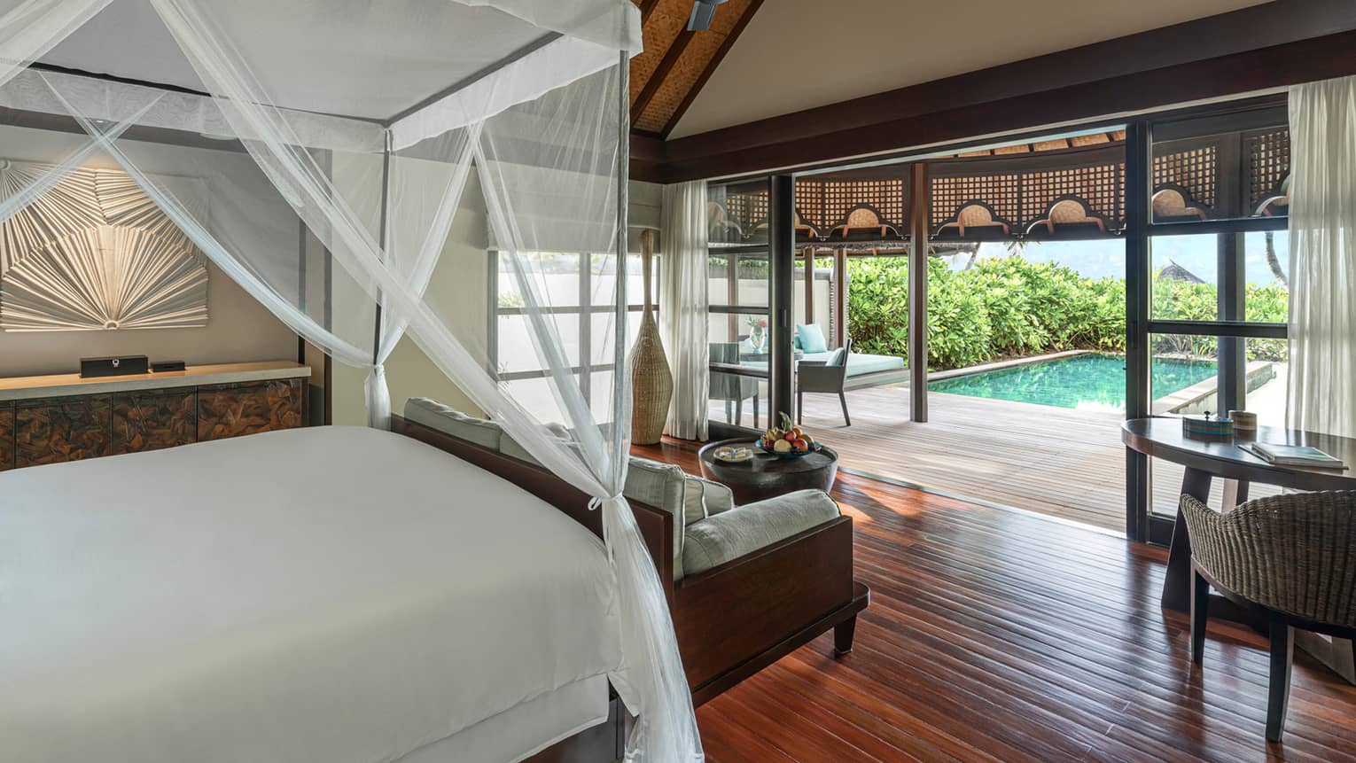 A white canopy hangs from the ceiling, covering a dark wood bed that overlooks a private pool surrounded by lush vegetation in the Sunset Beach Bungalow  
