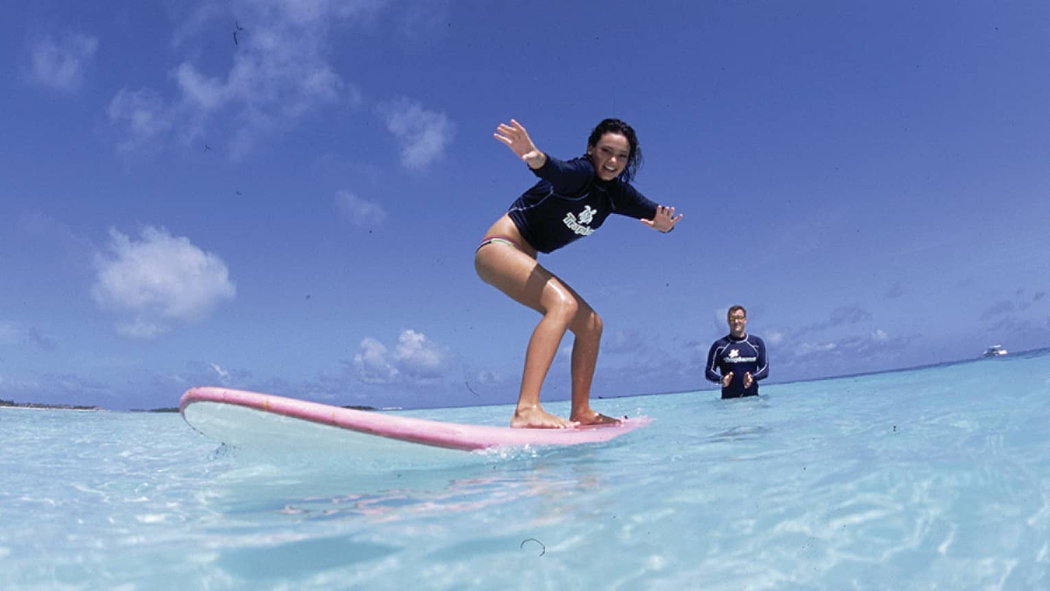 Woman rides pink surfboard on crystal clear water with man behind clapping