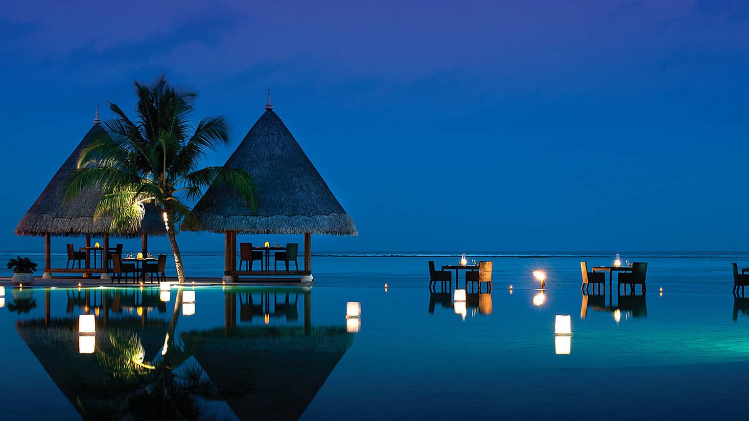 Kandu Grill at night, with candlelit dining tables under a towering palm, perched on ocean and infinity pool