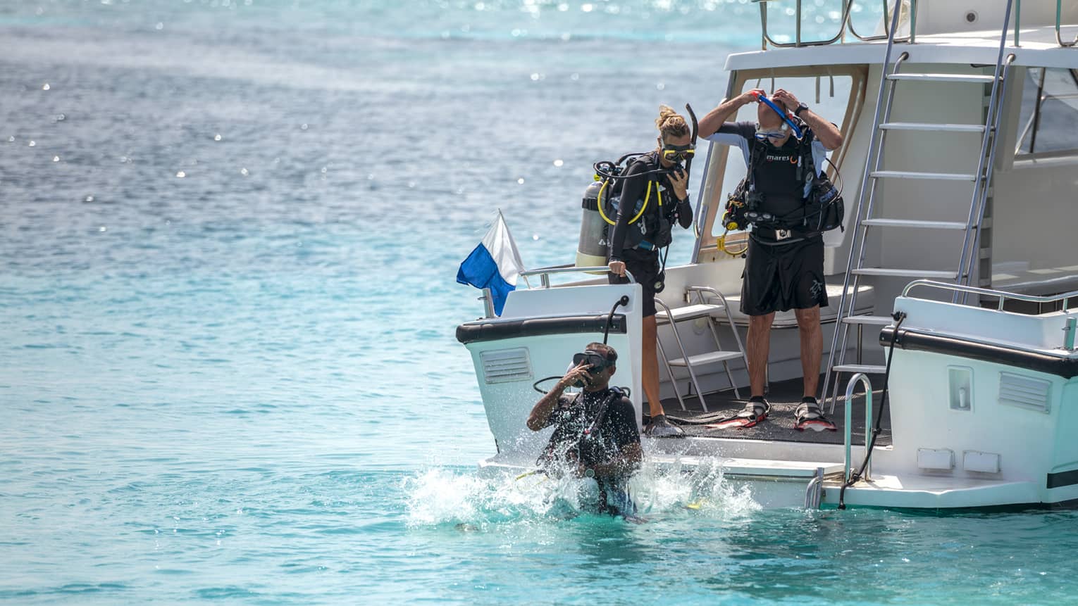 One scuba diver in water, two other divers stand on boat preparing to jump