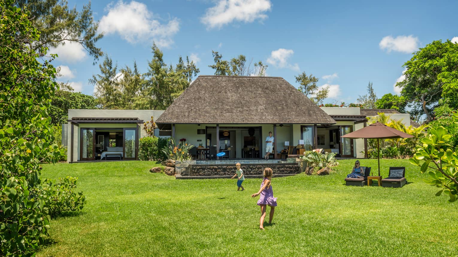 Young children play on green lawn in front of bungalow as dad watches from covered patio