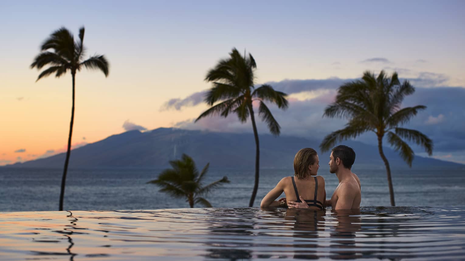 Man rests hand on woman's shoulder in infinity swimming pool, looking out at ocean, mountain and sunset