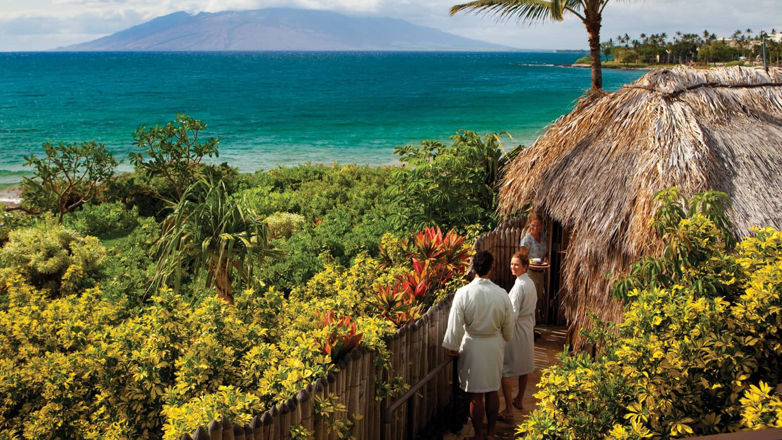 Couple in white bathrobes follow spa attendant into thatched-roof hut on path looking out at ocean