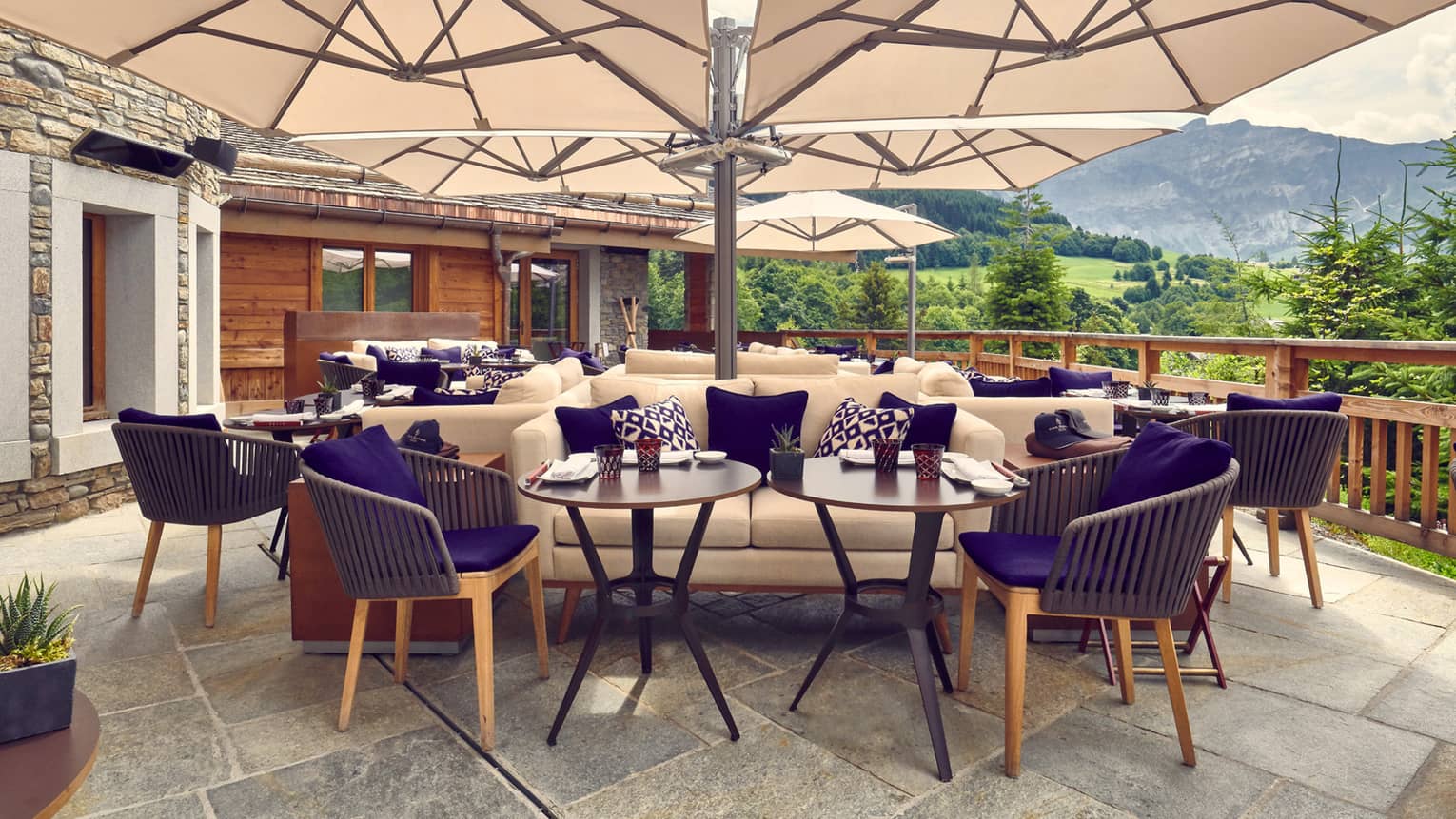 Restaurant flagstone terrace with tan and purple lounge seating under large tan umbrellas, mountain views behind