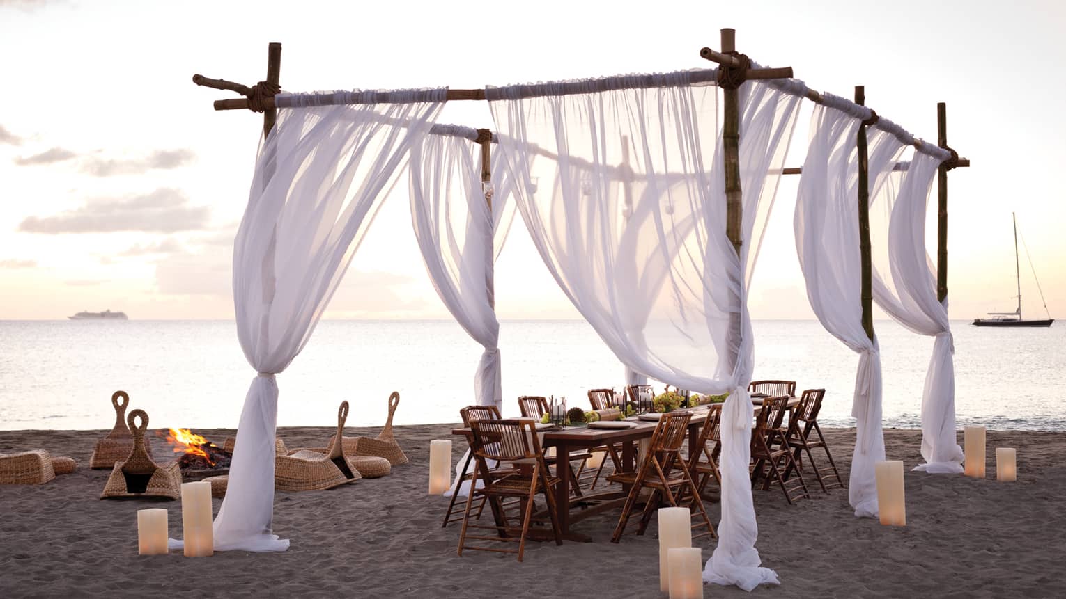 Sheer white curtains draped across wood posts around dining table on beach at sunset