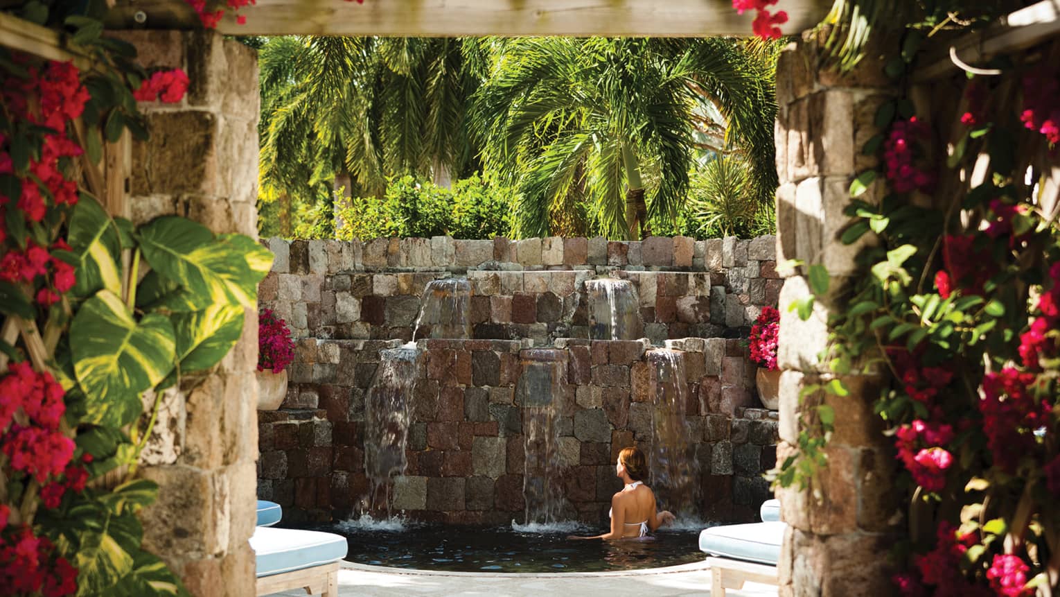 Woman bathes in small pool under stone fountain under palm trees, brick walls with pink flowers