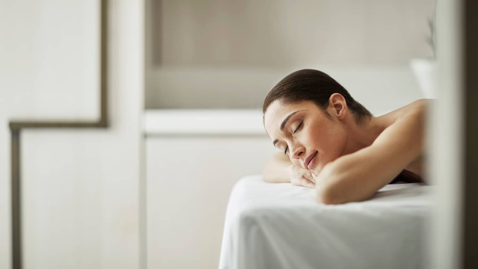 Woman smiles with eyes closed, rests head on arms as she lies on massage table with white sheet