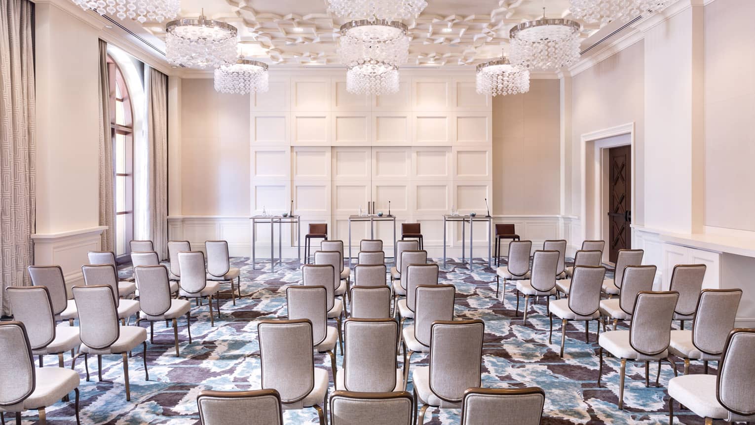 A cream colored function room is set up with rows of neutral chairs lined up and facing three tables in the front