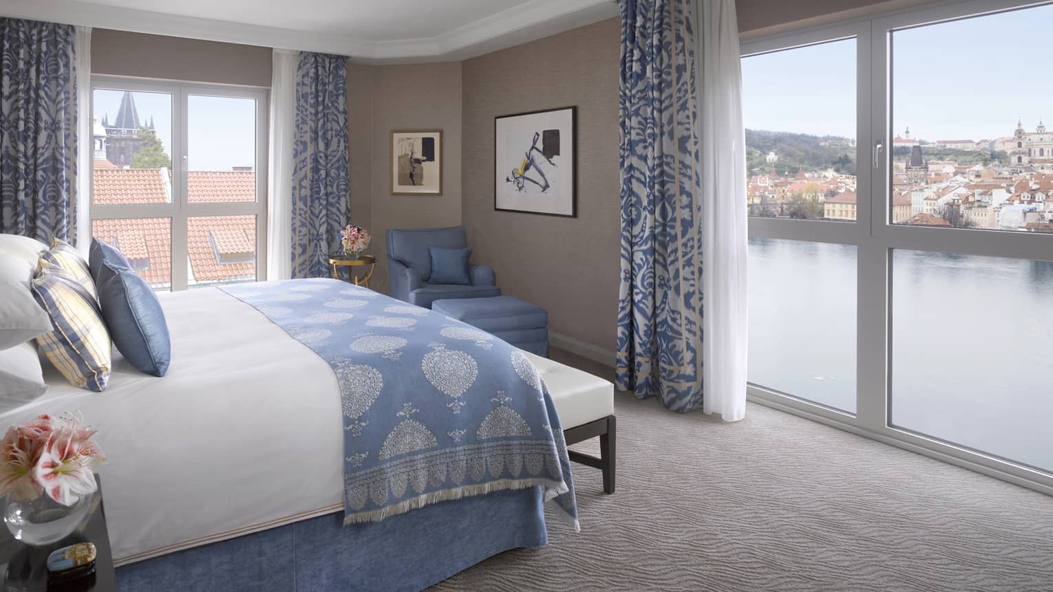 Premier Suite bed with blue blanket, pillow accents, in front of floor-to-ceiling corner windows overlooking Vltava River 