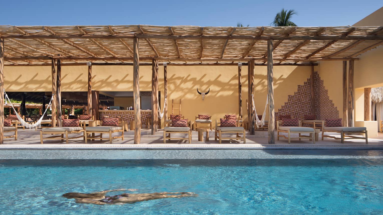 Woman swims in turquoise Tamai adults-only pool, lounge chairs under bamboo patio awning