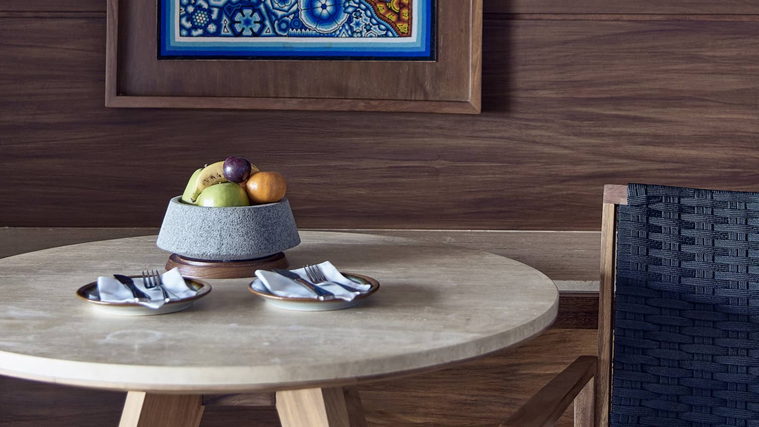 Circular table with bowl of fruit, arm chair, hanging artwork on wooden wall