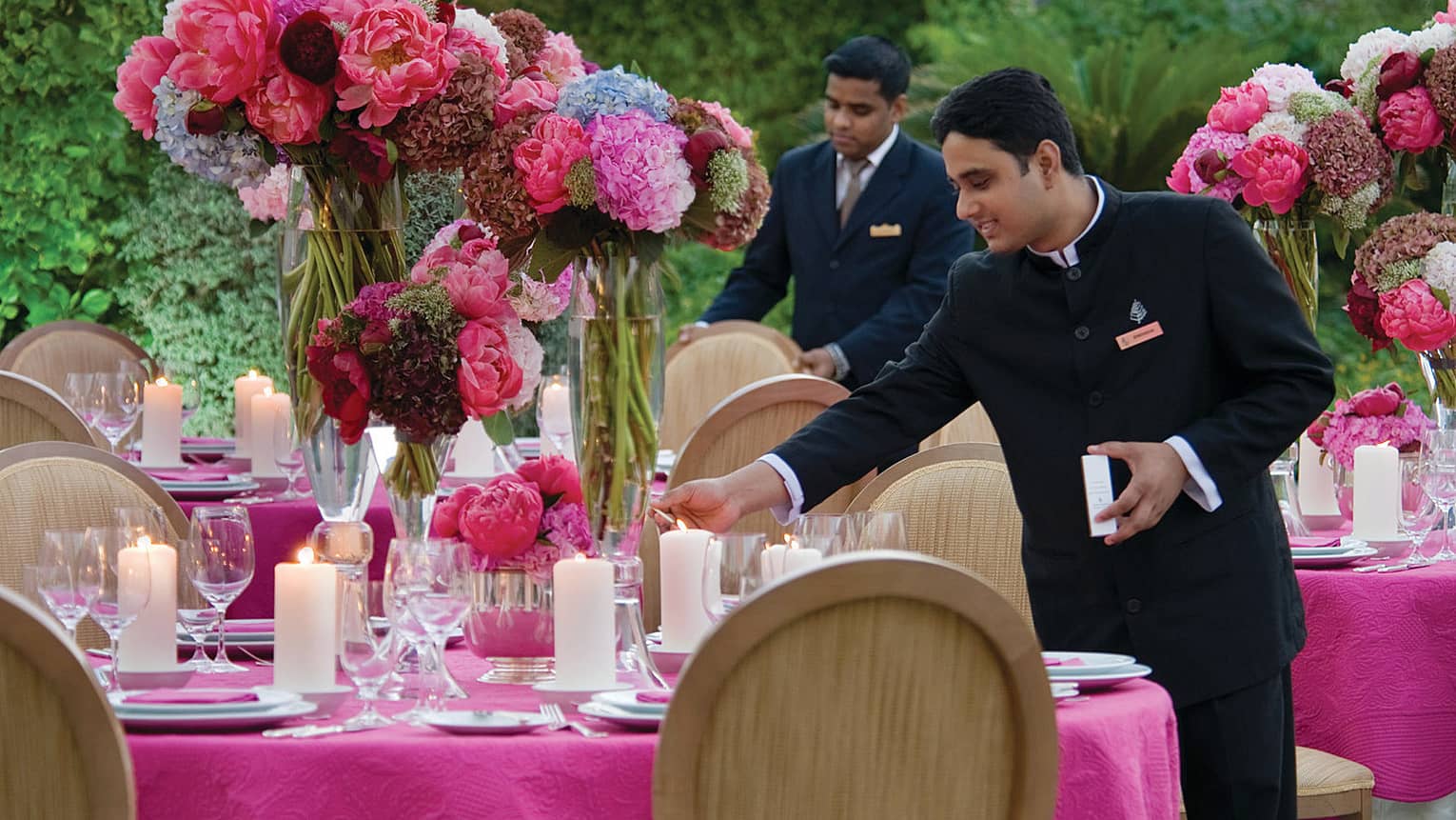 Two male staff set table with pink tablecloth, large vases of flowers and candles