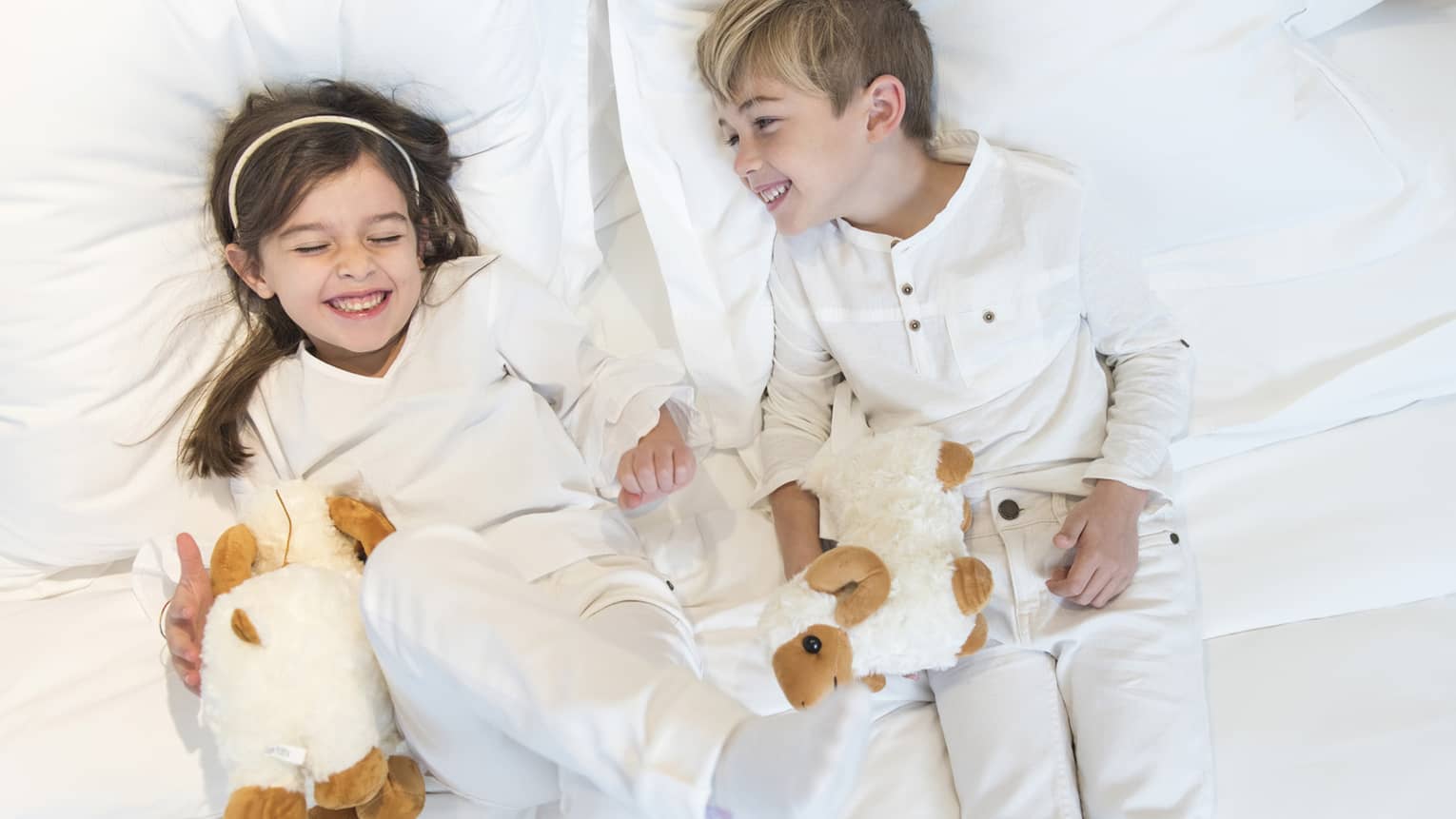 Aerial view of two children wearing white pyjamas with white stuffed animals lying on bed, laughing